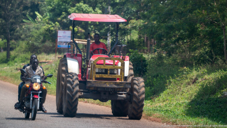 Red farm tractor and motorcyclist approaching together on a narrow paved road bordered by thick vegetation.