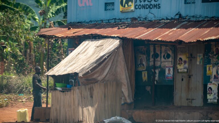 Small timber kiosk with a sloping metal roof in front of a weathered shop covered with posters and health advertising.