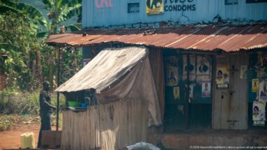 Small timber kiosk with a sloping metal roof in front of a weathered shop covered with posters and health advertising.