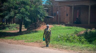 Woman in a patterned dress and head covering carrying a green container beside a brick building and grassy yard.