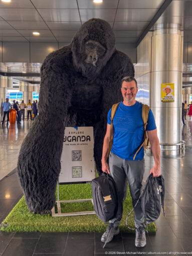 Traveler with bags posing beside a large gorilla sculpture and Explore Uganda display inside an airport terminal.