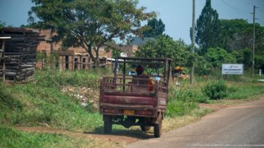 Red three-wheeled utility vehicle driving away on a paved road beside brick buildings, timber stacks, and greenery.