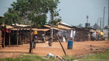 Cluster of timber roadside dwellings with residents, hanging laundry, cooking smoke, a blue barrel, and open dirt ground.