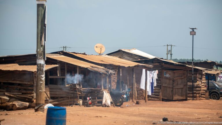 Timber roadside shacks with corrugated roofs, hanging laundry, smoke from an outdoor fire, and a blue plastic barrel.