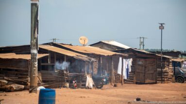 Timber roadside shacks with corrugated roofs, hanging laundry, smoke from an outdoor fire, and a blue plastic barrel.