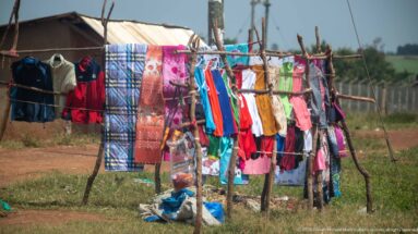 Colorful dresses, shirts, trousers, and textiles hanging from a rough timber rack beside a dirt roadside.
