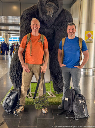 Two male travelers with backpacks and luggage posing beside a large gorilla sculpture inside an airport terminal.