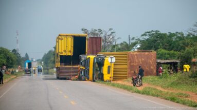 Distant roadside view of an overturned yellow truck, shipping containers, pedestrians, and a motorcyclist beside an open highway lane.