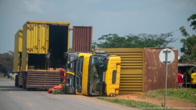 Overturned yellow truck beside stacked shipping containers and a flatbed trailer at the edge of a paved road.