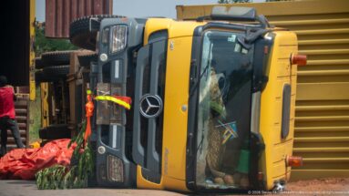 Yellow Mercedes truck overturned on its side beside the road with red material and spilled green produce nearby.