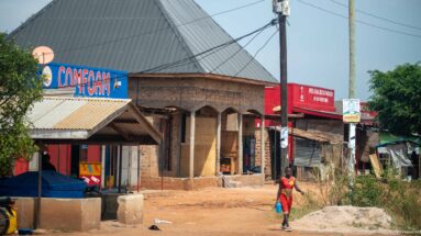 Campcom-branded roadside shop beside an unfinished brick building with arched openings, metal roof, and dusty frontage.