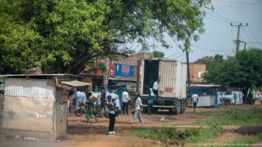 Box truck beside roadside businesses with workers, bicycles, sacks, corrugated structures, and a large shade tree.