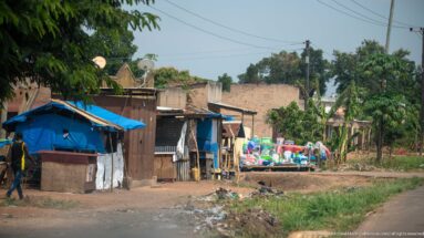 Timber, metal, and blue-tarp roadside structures beside brick buildings, merchandise, utility lines, and exposed earth.