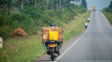 Motorcyclist carrying stacked crates uphill on a long rolling highway with other riders and a truck in the distance.