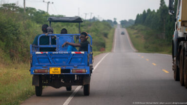 Blue three-wheeled cargo vehicle carrying several passengers along the shoulder of a rolling paved highway beside a truck.