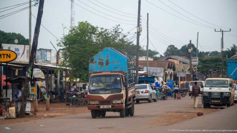 Isuzu cargo truck driving through a busy commercial street with cars, motorcycles, pedestrians, and roadside shops.