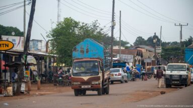 Isuzu cargo truck driving through a busy commercial street with cars, motorcycles, pedestrians, and roadside shops.