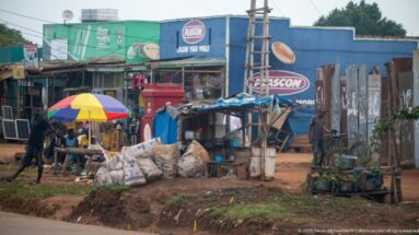 Informal stalls with sacks and tarps beneath a colorful umbrella in front of Kascon-branded roadside shops.