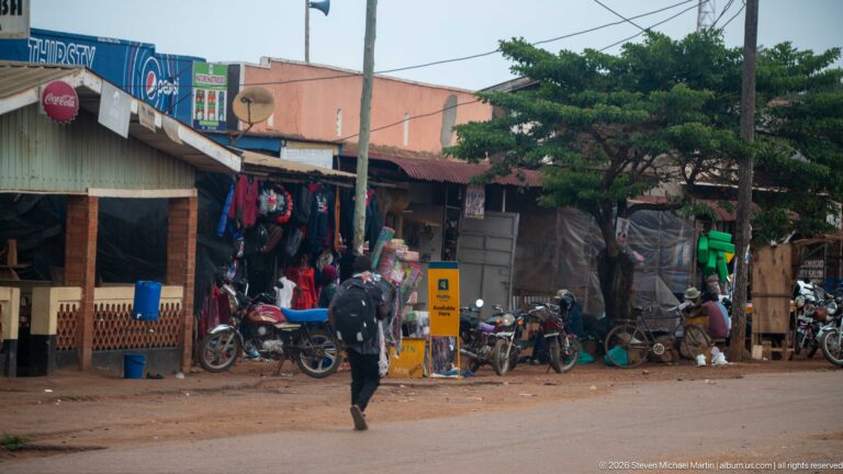 Pedestrian crossing a street lined with parked motorcycles, hanging clothes, and open-front roadside shops.