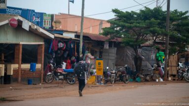 Pedestrian crossing a street lined with parked motorcycles, hanging clothes, and open-front roadside shops.