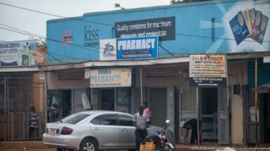 Azima Pharmacy storefront beneath a large blue condom advertisement with a parked silver car and motorcycle outside.