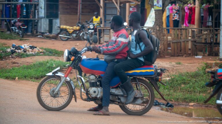 Two people riding a blue motorcycle past roadside shops, clothing displays, parked bikes, and a dirt verge.