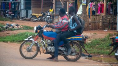 Two people riding a blue motorcycle past roadside shops, clothing displays, parked bikes, and a dirt verge.