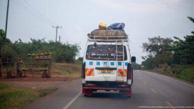 Rear view of a white minibus carrying mattresses, sacks, and bulky goods on its roof along a paved highway.