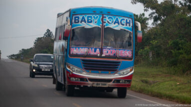 Blue and red Baby Coach bus marked Kampala Express approaching on a two-lane Ugandan road.