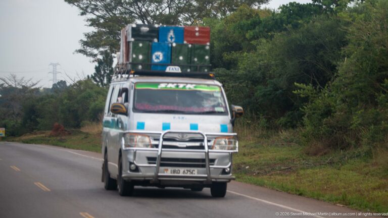 Passenger minibus approaching on a paved road with colorful suitcases and luggage stacked high on its roof rack.