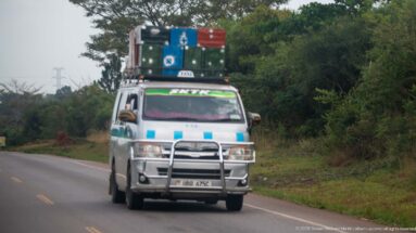 Passenger minibus approaching on a paved road with colorful suitcases and luggage stacked high on its roof rack.