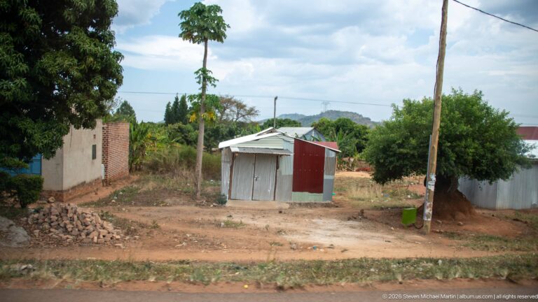 Small corrugated-metal kiosk with a pitched roof on a cleared dirt plot between homes, vegetation, and a pile of stones.