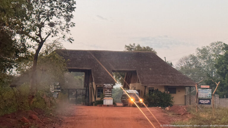 Vehicle with headlights approaching a thatched entrance or reception structure on a red dirt road at dusk.