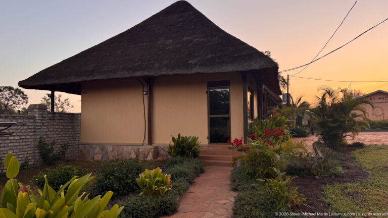 Small round lodge bungalow with steep thatched roof, garden path, flowering shrubs, and warm sunset sky.