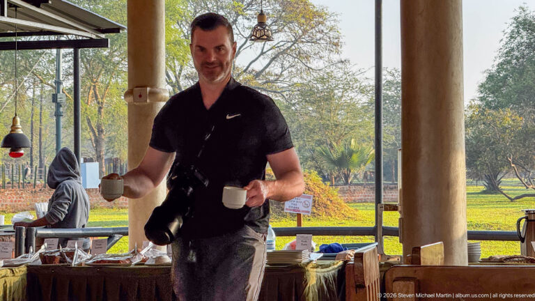 Man carrying two mugs with a camera at his side inside an open-sided lodge dining or service area.