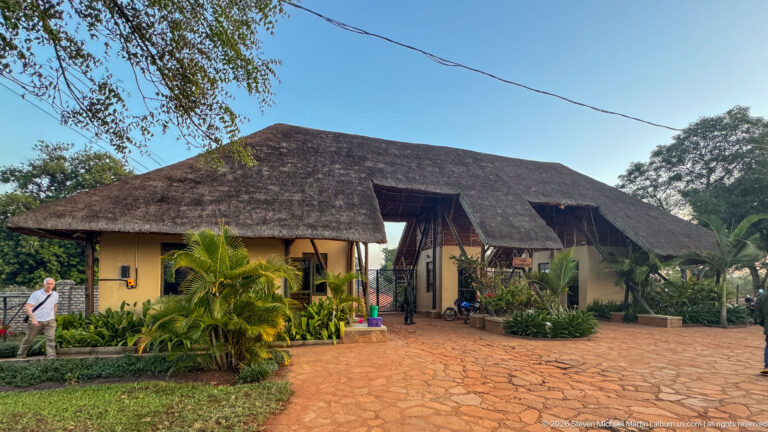 Wide thatched-roof lodge building with open central entrance, stone-paved forecourt, and dense tropical landscaping.