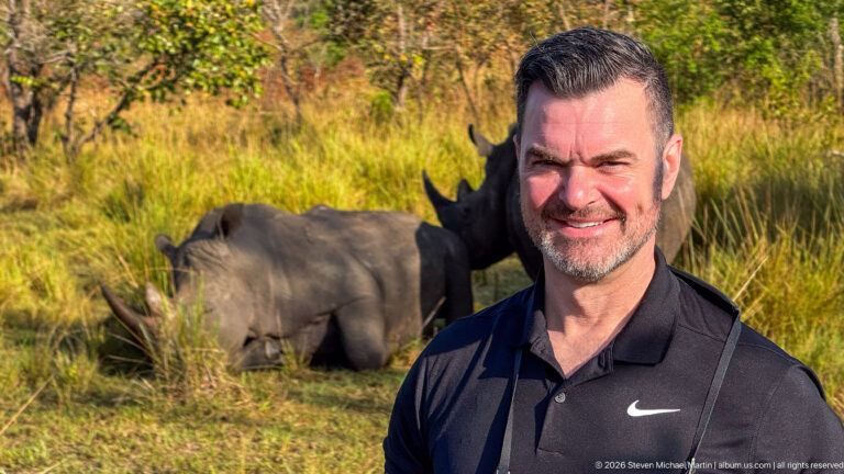 Smiling man in black polo in foreground with two white rhinoceroses resting in sunlit grass behind him.