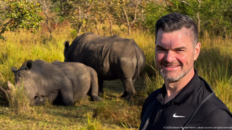 Smiling man in black shirt in foreground with two white rhinoceroses resting in tall grass behind him.