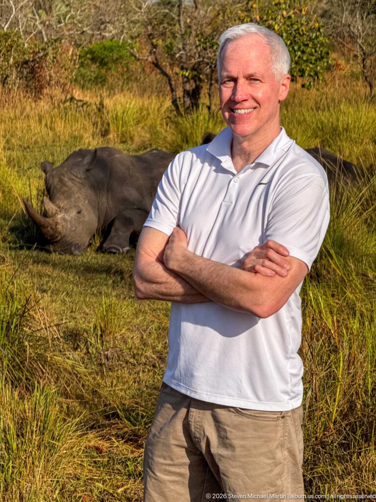 Smiling man in a white polo standing in front of resting white rhinoceroses in sunlit grass.