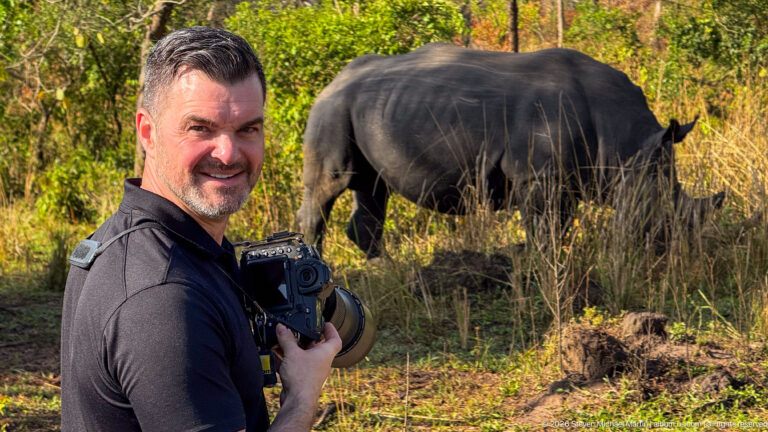 Man holding a camera in the foreground with a white rhinoceros grazing behind him among dry grass and trees.
