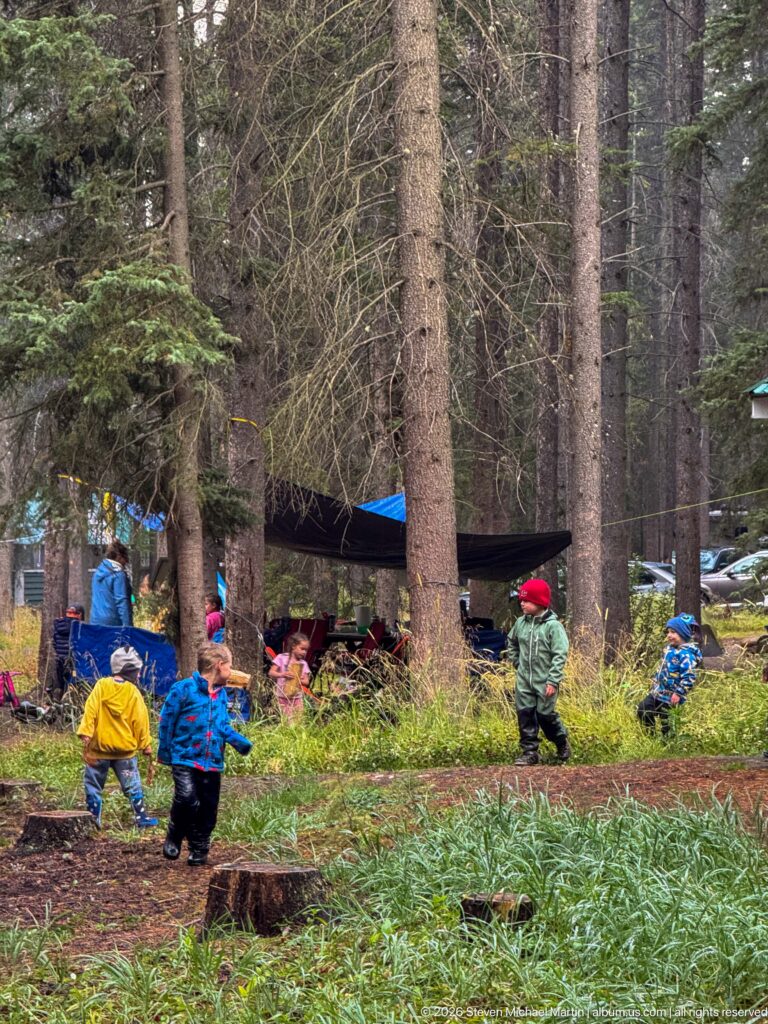 Children and adults gather near a canopy and tents in a wooded campground clearing.