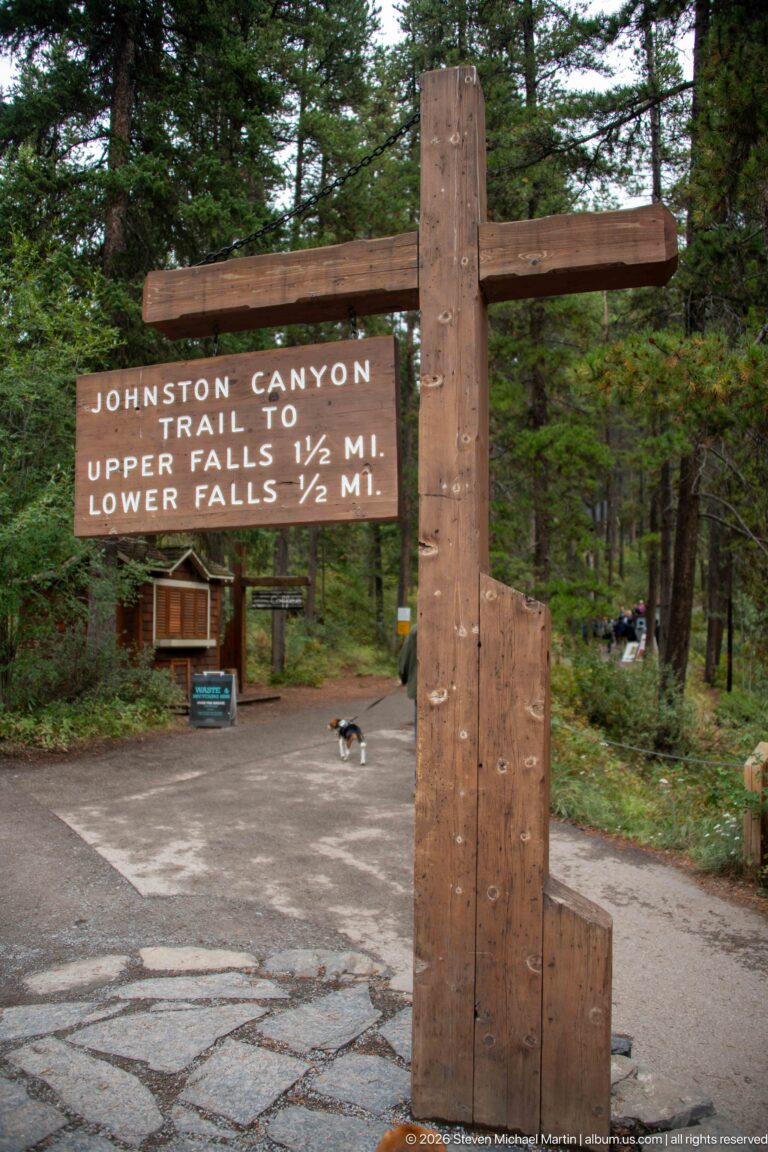 Wooden trail sign lists distances to Upper and Lower Falls beside a forested paved approach.