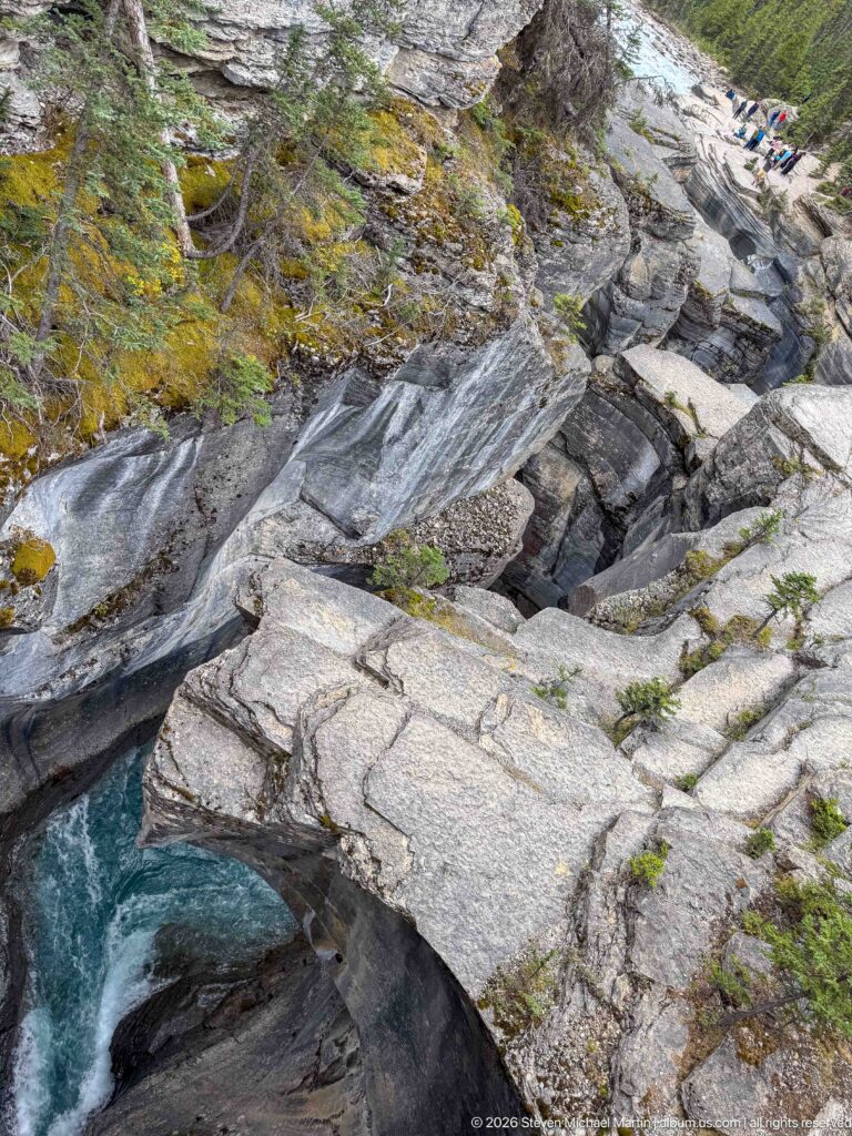 Fractured pale rock slabs slope toward a narrow turquoise channel, with visitors visible above.