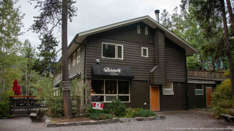 Dark two-story timber building with a storefront awning, Canadian flag, and outdoor seating among conifers.