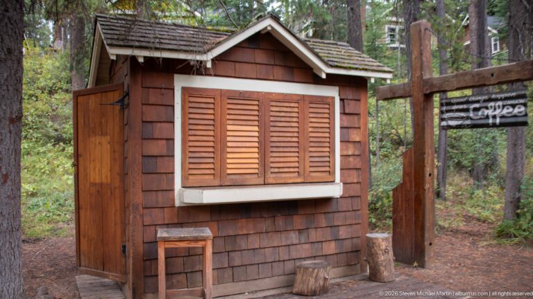 Small timber coffee kiosk with closed shutters, counter, stool, and a hanging “Coffee” sign among trees.