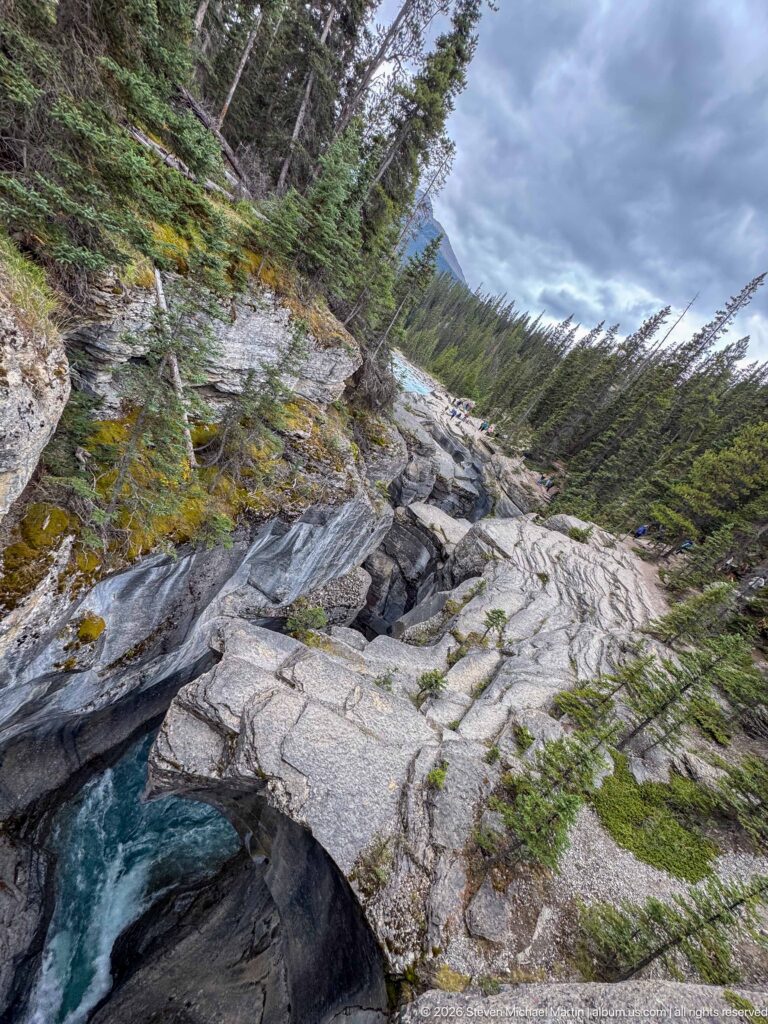 Exposed rock apron descends toward a creek beneath forested canyon slopes, with visitors in the distance.