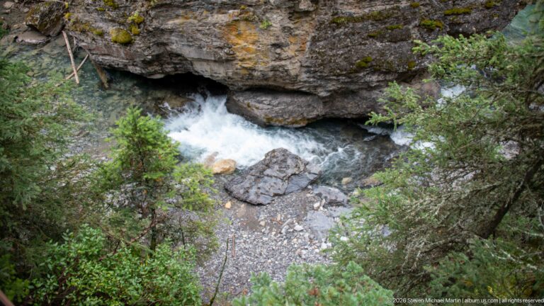 Small waterfall drops beneath a projecting rock ledge into a shaded rocky basin.