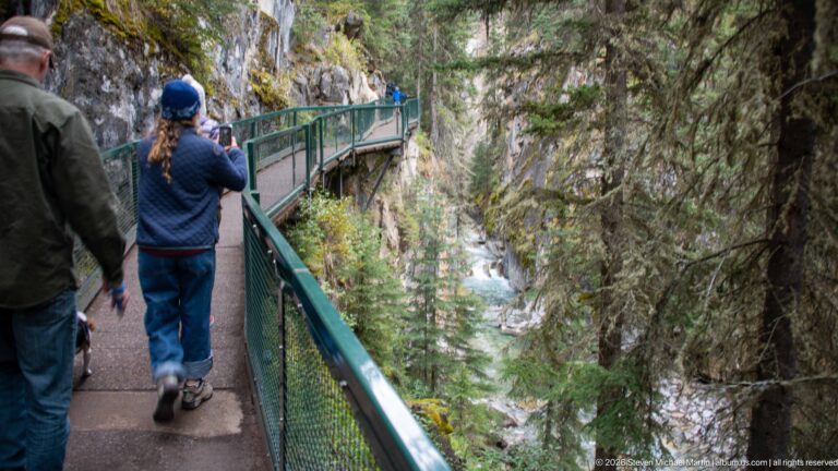 Two hikers walk a green-railed boardwalk above the creek; one raises a camera toward the canyon.