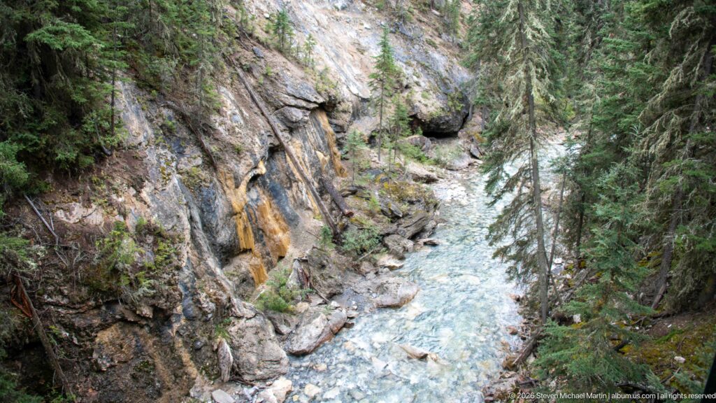 Rocky stream winds through a conifer-lined canyon beneath stained rock walls and forested slopes.