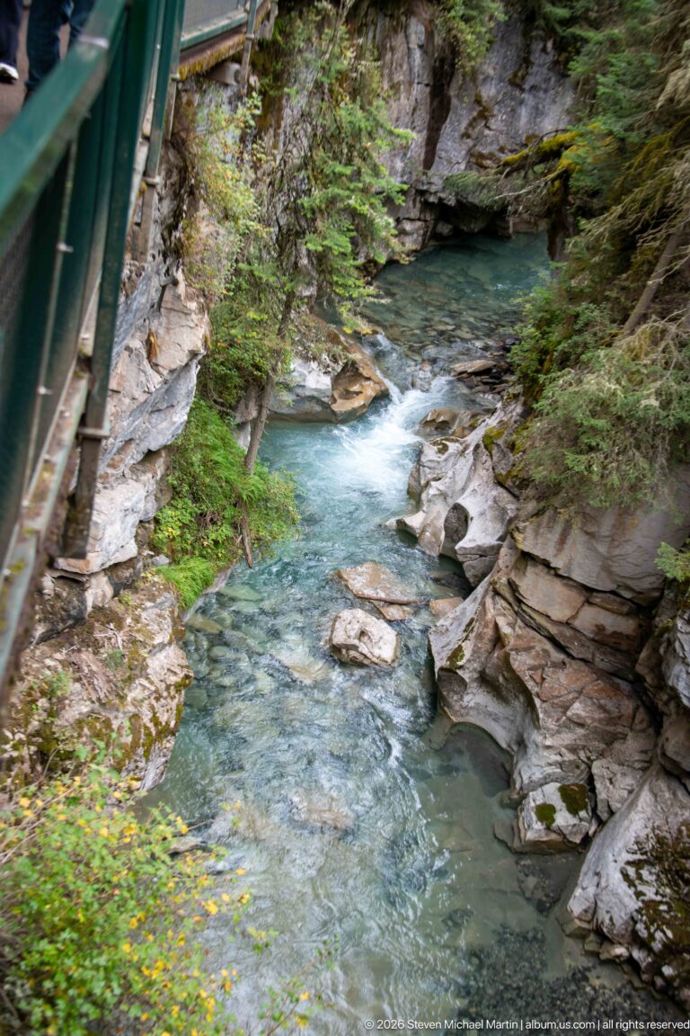 Green-railed walkway borders a rocky creek channel with small cascades and a blue-green pool.